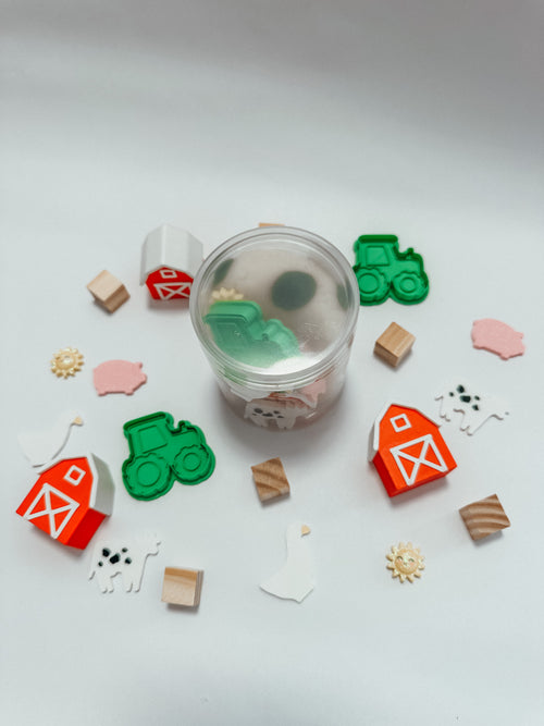 A clear plastic jar filled with small farm-themed pieces, surrounded by scattered matching pieces including green tractors, red-and-white barns, wooden blocks, white pigs, sheep, and sun shapes on a light gray background.