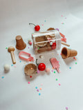 A miniature toy ice cream set arranged on a white background, including a clear cylindrical container with toy waffle cones and cherries, small pink popsicle pieces, brown cone-shaped pieces, a wooden scoop, and scattered pastel confetti.