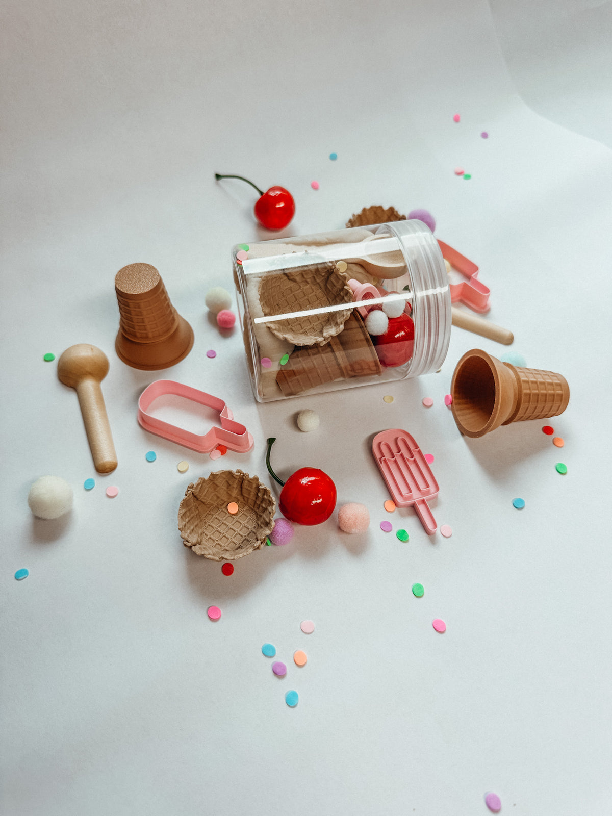 A miniature toy ice cream set arranged on a white background, including a clear cylindrical container with toy waffle cones and cherries, small pink popsicle pieces, brown cone-shaped pieces, a wooden scoop, and scattered pastel confetti.