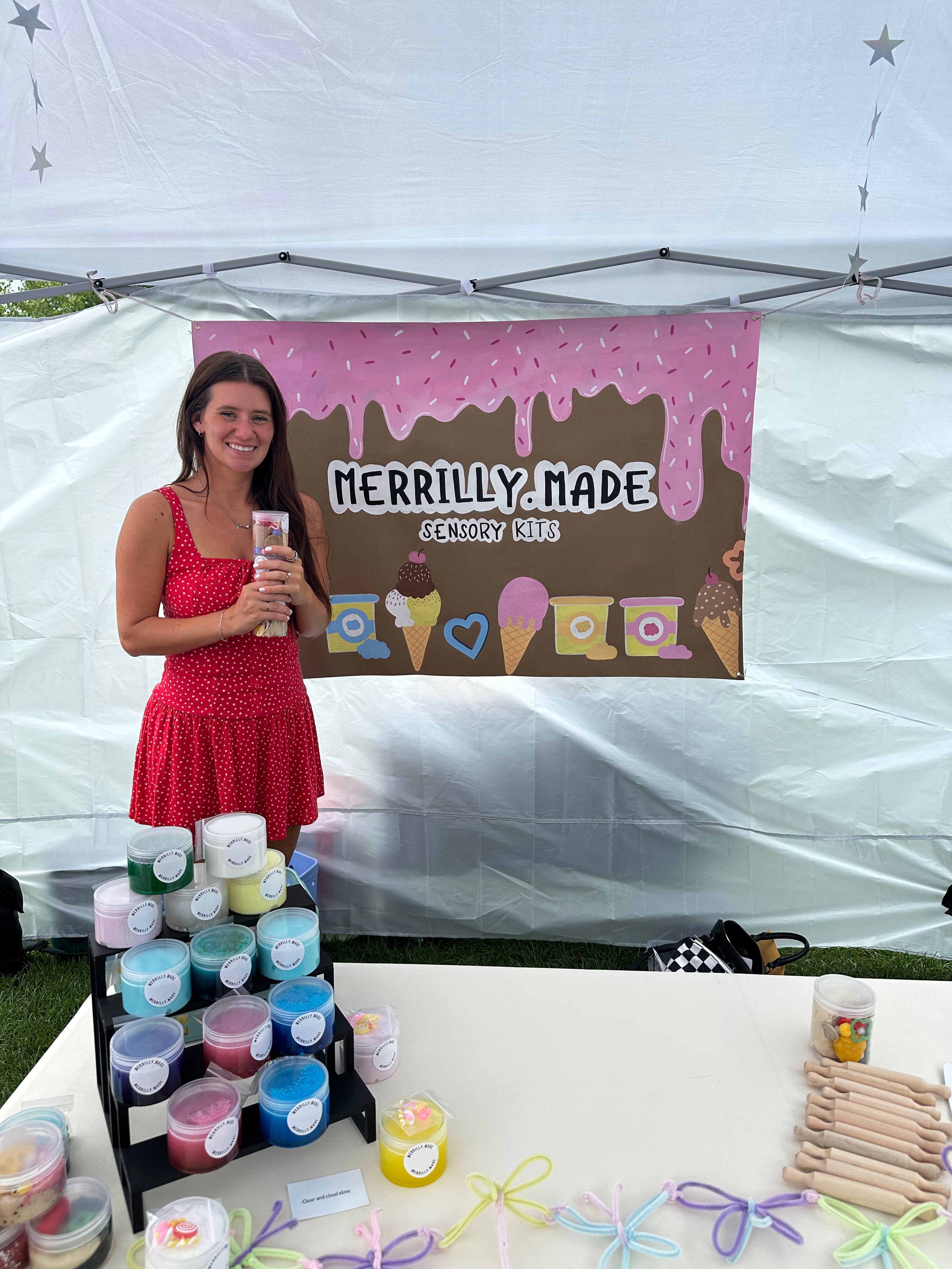 A woman in a red dress stands behind a table displaying colorful candle jars and wooden wicks under a white canopy tent. A large banner behind her reads 'MERRILLY MADE ENGLAND Candles' with pink and brown graphics.