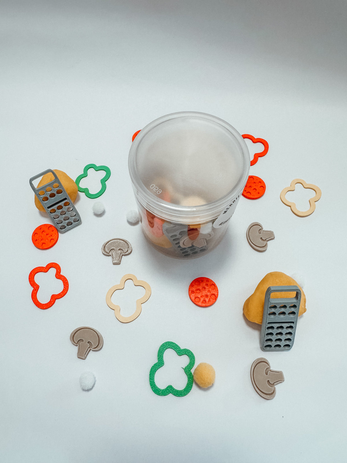 A clear plastic container filled with small colorful food-themed pieces, surrounded by loose matching pieces including mushroom shapes, tomato circles, leaf shapes, and grater pieces on a white background.