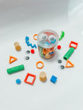 A clear plastic jar filled with colorful craft and sensory pieces, surrounded by loose matching pieces on a white studio background. The set includes googly eyes, orange rings, blue and green wavy pieces, red square frames, tan cylinders, and other bright plastic shapes.