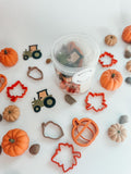 A clear plastic jar filled with small decorative fall-themed pieces, surrounded by orange and beige pumpkins, leaf-shaped cookie cutters, tractor cutters, and acorn cutters on a white background.
