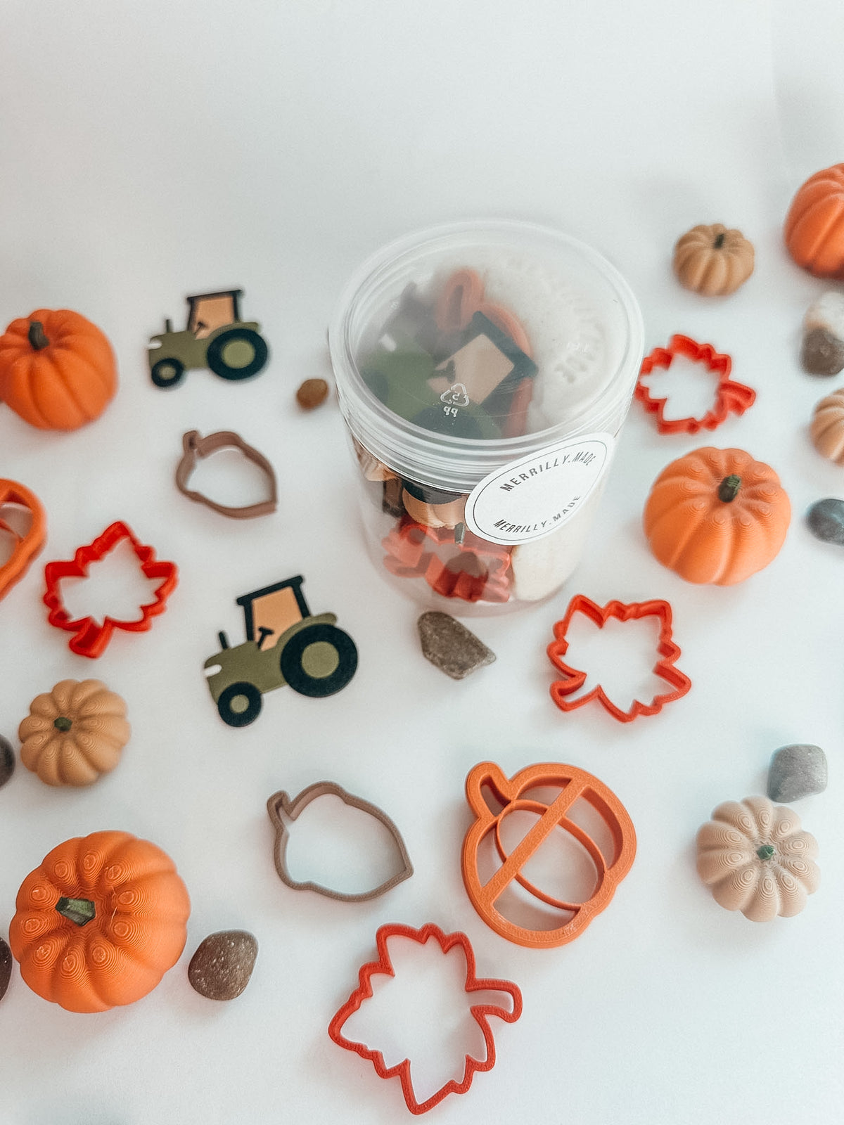 A clear plastic jar filled with small decorative fall-themed pieces, surrounded by orange and beige pumpkins, leaf-shaped cookie cutters, tractor cutters, and acorn cutters on a white background.